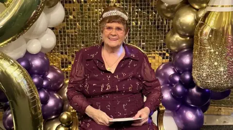 Supplied A picture of an elderly lady sat down wearing a purple dress,glasses and a tiara with 90th birthday balloons behind her.