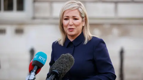PA Media Michelle O'Neill stands in front of three microphones before a white brick building. She has shoulder-length blonde hair and is wearing a navy coat.