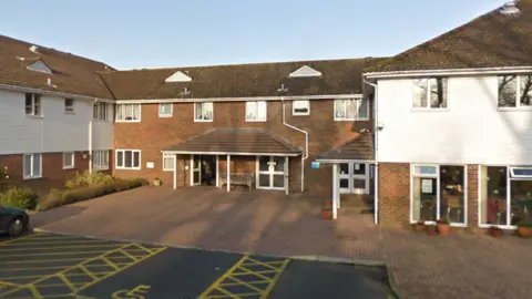 Google The entrance to Ticehurst Surgery, a collection of brown brick two-storey buildings with white cladding to the left and right and spaces for disabled parking opposite the main door. An elderly man can be seen leaving the building