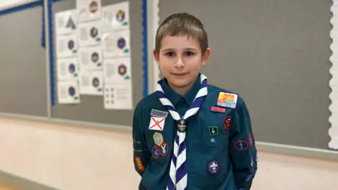 BBC Charlie looks at the camera, he is wearing his scouts uniform which is a dark green shirt with a blue and white neckerchief. He has several bright badges sown onto his uniform. Charlie has dark, brown eyes and short light brown hair.