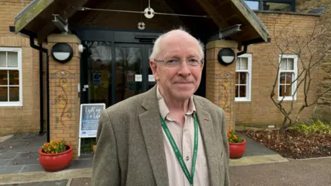 St Cuthbert's Hospice Paul Marriott standing in front of the entrance to St Cuthbert's Hospice. He is bald with short grey hair at the sides and is wearing glasses and a tweed blazer and khaki shirt. The building behind him is made from brick and has glass doors, tinted black.