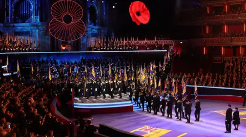 PA Media Standard bearers carry flags in the Royal Albert Hall during last year's Festival of Remembrance, with two large poppies above. Hundreds of people watch in the audience.