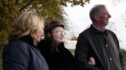 BBC Emily in centre has brown hair and is wearing glasses and a black coat, with her parents. Her mum has blonde hair and is wearing a blue jacket, her dad is has short hair and is wearing a brown jacket.