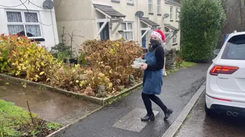 Susan Clarke Sue Clarke outside of a house wearing a red Christmas hat with a blue pinafore and black. She is walking towards a house with Christmas meals in her hands. The garden has bushes and flowers in the front. 