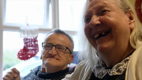 BBC Barry has glasses and short grey hair. He is sitting next to his fiancée Betty who is grinning. There is a red and white sparkly Christmas stocking hanging on the window next to them