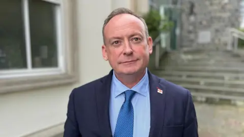 BBC Deputy Lyndon Trott looking towards the camera, he has brown hair with flecks of grey and is wearing a blue suit with a Guernsey pin badge, with a blue shirt and metallic blue tie.
