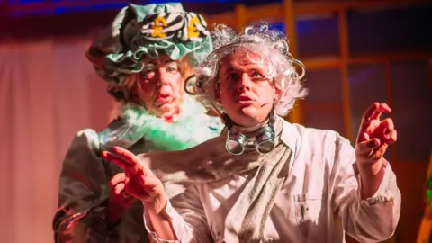 BBC Two male actors on stage. The one in front is wearing a beige shirt, scarf and hat. He has unruly white, curly, hair and has large goggles around his neck. He is gesticulating with his hands. The man behind him is dressed as a  pantomime dame. He is wearing a dress and has green feathers around his neck and a green bonnet on his head featuring yellow pound signs.