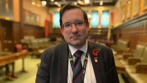 Lawrie Hooper, who has dark hair and is standing in the House of Keys Chamber, wearing glasses, a poppy, a striped tie and an Isle of Man TT lanyard.