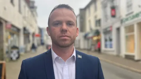 A man with a short beard standing in a shopping street looking straight at the camera wearing a blue jacket, white shirt and Cornish flag pin badge