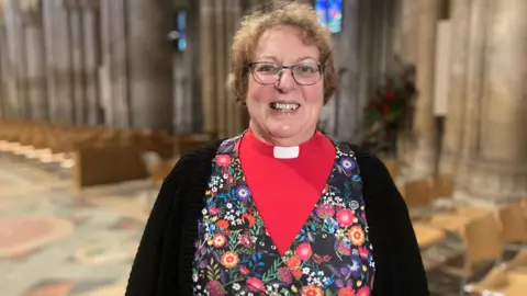 Jenny Kirk/BBC Rev Canon Fiona Brampton stands in Ely Cathedral facing the camera and smiling broadly. She is wearing a bright red shirt with a white dog collar, and over that a flowery patterned top and then a black cardigan. She has green glasses and short light brown curly hair. Chairs and pews are in the background, not in focus. 