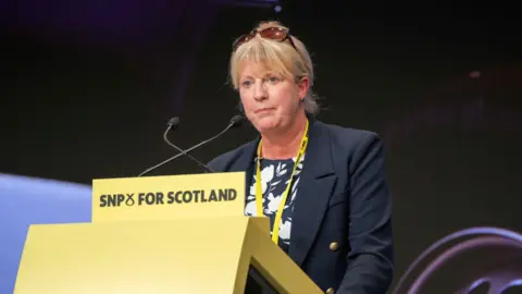 PA Media Shona Robison, with glasses resting on top of her head and dressed formally, making a speech at an SNP conference. A yellow conference lanyard is around her neck and the yellow podium she is standing behind says SNP FOR SCOTLAND on it