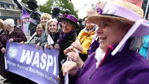 Getty Images Women in bright purple clothing stand in a line behind a Waspi campaign sign  