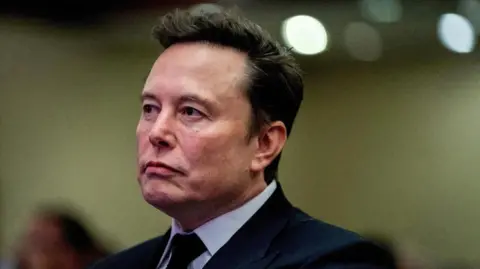 Reuters Elon Musk is looking away from the camera. He is wearing a dark coloured suit, white shirt and dark coloured tie.