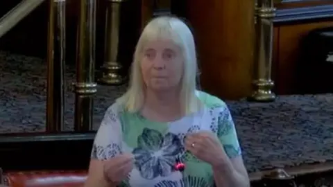 Margaret Aspinall A woman with white blonde hair and a fringe is smiling. She is wearing a white top with green spots and black flowers on it. 