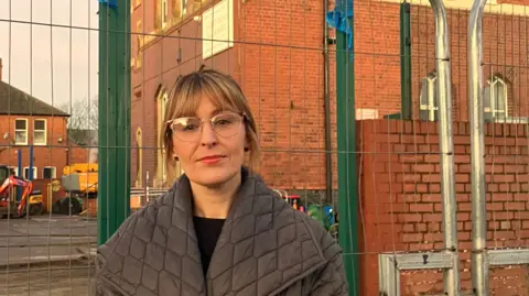 BBC Simone Beach is standing in front of the red-brick two-storey brick building. She has blonde hair and is wearing glasses and a grey coat.