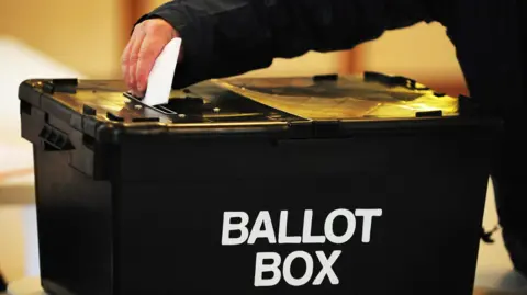 PA Media A black ballot box with a hand seen above it putting a folded sheet of paper through a slot in the top. On the side of the box the words ballot box are written in large, white capital letters.