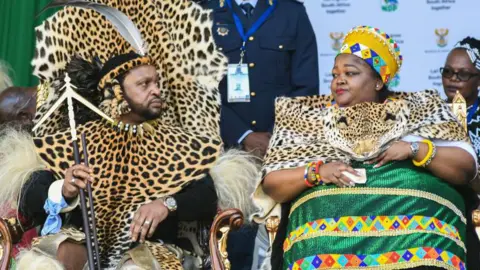 Getty Images South Africa's Zulu king and his first wife are seen in colourful traditional attire during an official event. The king is wearing a leopard print wrap over his black long sleeved top while the queen is also wearing a similar wrap over her traditional green dress.
