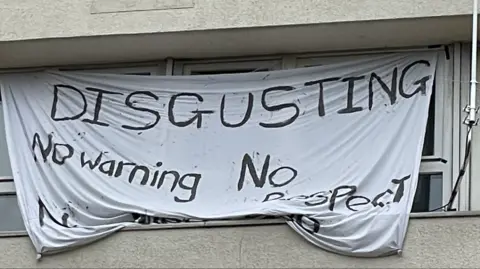 BBC A white sheet hung up across a window in Barton House in Bristol, with painted words reading: "Disgusting, no warning, no respect"