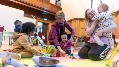 PA Media Nicola Sturgeon playing with children at a day care event - she has a face mask on and is is wearing a purple jacket. She is sitting down. Two children are also sitting down and playing with toys on the ground. Another woman, with glasses, blonde hair and a pink top, is also sitting down and is talking to another child, in her arms.