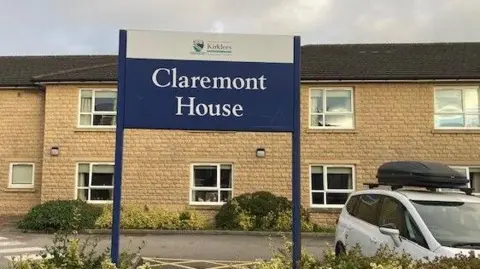 BBC Sign saying 'Claremont House' in forefront of picture, with a two-storey sandy brick building in the background. There is a white car parked in the car park to the right of the picture.
