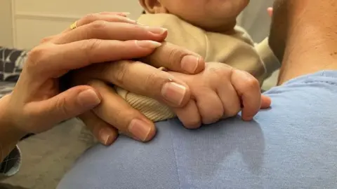 Image of a man taken from behind over the shoulder holding the hand of a baby. A separate female hand is placed on top of the man's hand