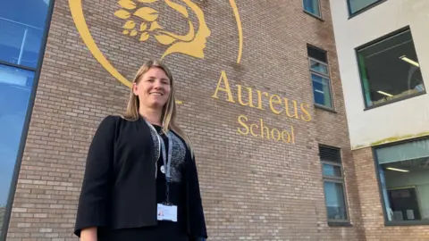 BBC Headteacher of Aureus School, Kirsty Rogers, poses, smiling, in front of the main school building which displays  a large logo of the school reading 'Aureus School Didcot'