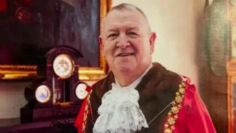 Bristol City Council Jeff Lovell wearing his red mayoral robes with fake fur trim and a white lace jabot. He is wearing gold mayoral chains and is smiling at the camera. HE is standing in front of a fireplace with a mantle clock and a large gold framed painting above it.