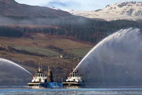 Royal Navy HMS Triumph submarine at sea, with two tugboats sailing alongside with spraying water cannon. Hills and trees are visible in the background.