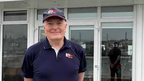 Man in cap and blue polo shirt smiles at camera standing in front of Guernsey Yacht Club
