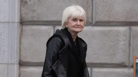 PA Media A woman with grey hair, wearing a black coat. She is looking at the camera and standing in front of a grey brick wall.