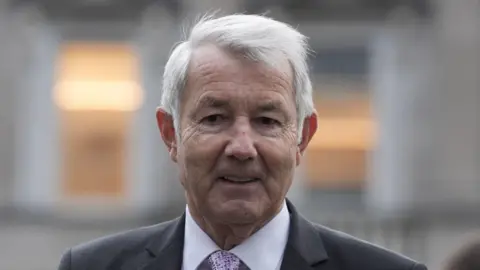 PA Media A man with grey hair, pictured from the shoulders up, wearing a grey suit and lilac shirt. The background of the image is blurred.