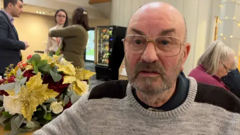 BBC A bald man in glasses with a short grey and brown beard, wearing a grey sweater with brown shoulder coverings, looks slightly to the left of camera. Behind him over his right shoulder are three members of staff, a man and two women, in conversation, with a flowery table decoration in the foreground. Behind the man's left shoulder is a seated woman looking down. She has grey hair in a pony tail.