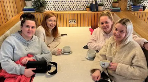 BBC/Julia Lewis Four young women are sitting around a table in a cafe booth. They each have a cup and saucer.