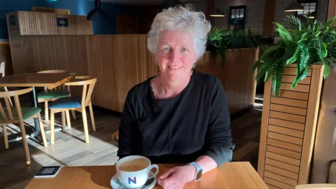 BBC/Julia Lewis A woman with short grey hair and wearing a black top and grey watch is sitting at a table with a cup of coffee in a white cup. There is another table and chairs behind the woman.