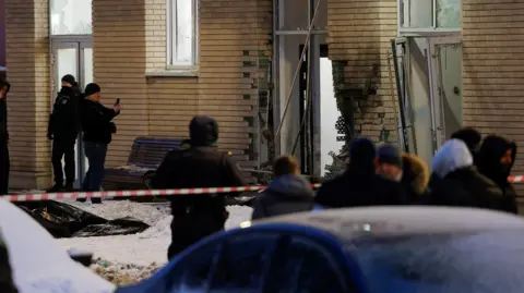 Reuters Scene of the explosion in which Igor Kirillov was reported killed. A damaged wall shows blackened and missing bricks, while two armed guards stand to the left and a crowd looks on from behind a red-and-white striped security cordon.