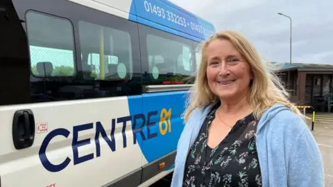 Andrew Turner/BBC Alison Holmes, wearing a patterned black top, and blue cardigan, has long blonde hair. She is smiling and standing next to one of the charity's fleet of minibuses which is adapted for people with physical and learning disabilities.