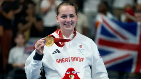 PA Tully Kearney, a young woman with scraped-back brown hair, smiles as she holds up a gold medal. She is wearing a white Great Britain Paralympics GB jacket.