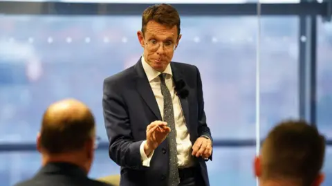 Jacob King/PA Wire A man stands up at a meeting to speak to attendees. He is wearing a dark blue suit, a white shirt, and a patterned tie. He has brown hair and glasses. The heads of two men attending the meeting can be seen from the back.