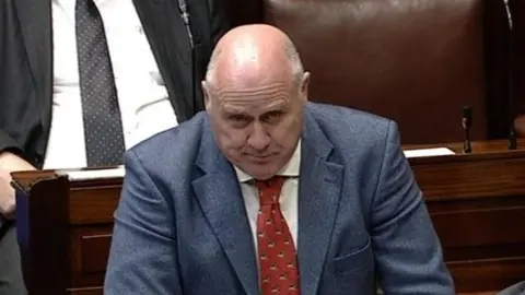 Oireachtas Noel Graelish speaking in the Dáil. He is bald and wearing a blue blazer, white shirt and red tie. The brown leather Dáil seats can be seen in the background with two other TDs sitting in them.