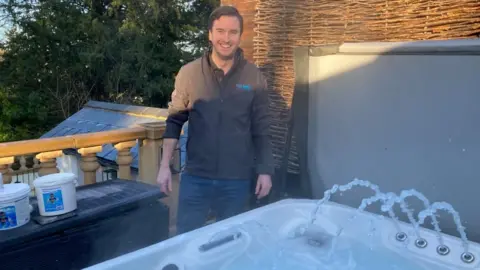 BBC James Symons standing behind of a hot tub with jets of water shooting in the air. He is wearing a brown jumper and smiling at the camera