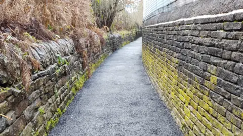 AIRE RIVERS TRUST A freshly-laid blue-black tarmac footpath with stone walls either side coverage in yellow and green moss.