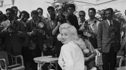 Getty Images Black and white image of Diana Dors wearing a white blouse, with a large number of photographers behind her.