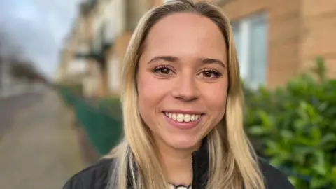 BBC Phoebe is stood in a street, smiling at the camera, she has long blonde hair and is wearing a black coat.