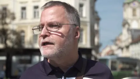 A man wearing spectacles is photographed outdoors in a city scene, though the background is blurred and difficult to see clearly