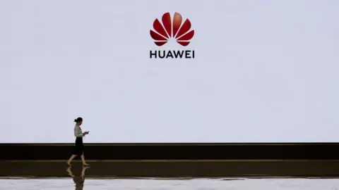 Getty Images A member of Huawei's reception staff walks in front of a large screen displaying the logo