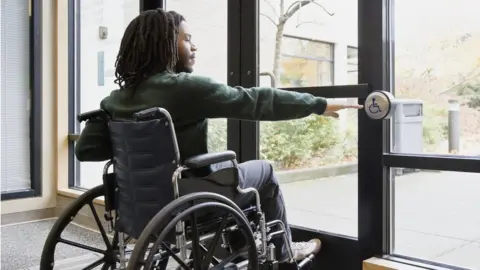 Getty Images Stock image of a wheelchair user entering a building using a door access button