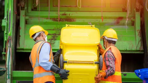 Getty Images Two refuge collectors load waste into the back of a green bin lorry from a bright yellow wheelie bin
