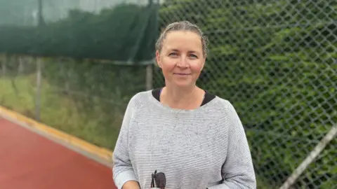 A white woman in sportswear standing by a tennis court fence