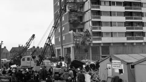 PA Media A black and white photo showing multiple collapsed floors and walls on the corner of Ronan Point, with cranes stretching into the sky on the left. Crowds of people stand in front of a large pile rubble on the ground