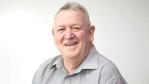 Bristol City Council Jeff Lovell wearing a light grey shirt and smiling at the camera. He has short white hair with dark grey patches, and is sitting in front of a white background.